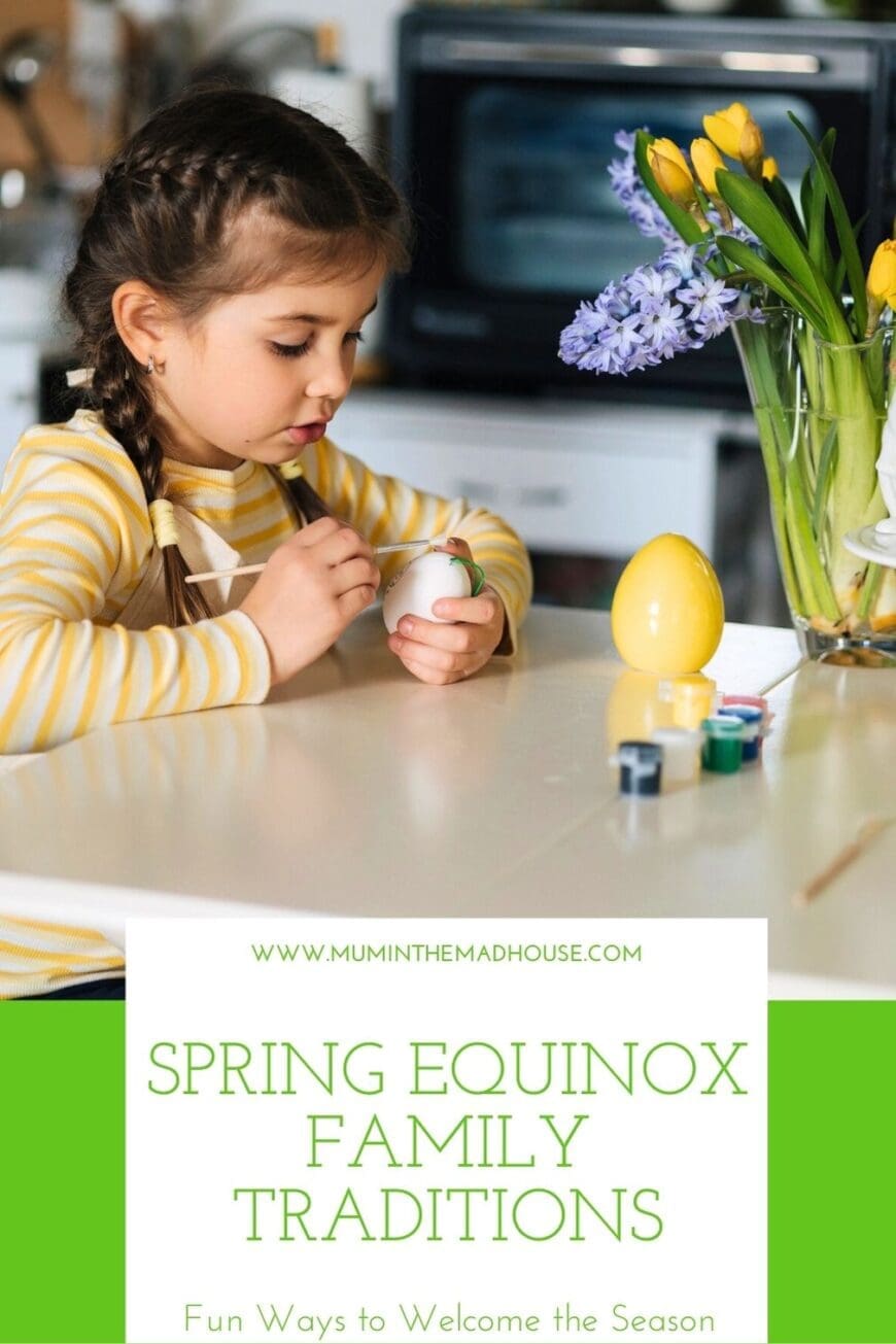 A little girl with braided brown hair, wearing a striped yellow and white shirt and an apron, is seated at a white kitchen table painting an egg for a spring-themed craft. A vase of fresh yellow daffodils and purple hyacinths sits on the table, alongside a collection of small paint pots and brushes. The text above reads: “Spring Equinox Family Traditions – Fun Ways to Welcome the Season” with the website www.muminthemadhouse.com displayed at the top.